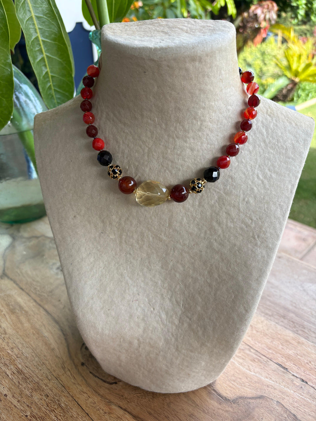 Collar corto artesanal de piedras semipreciosas en tonos rojos, negros y dorados exhibido en busto crema en la tienda.